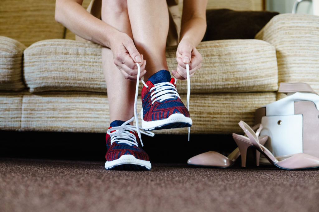 Woman lacing up running shoes next to dress heels and purse on the floor. 